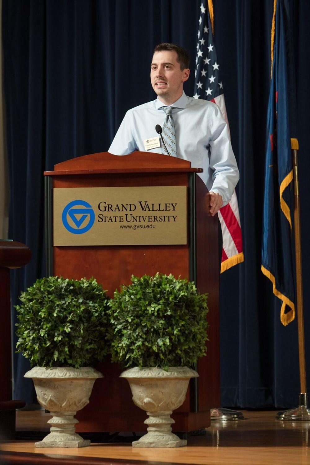 A man standing at a GVSU podium, speaking to the audience